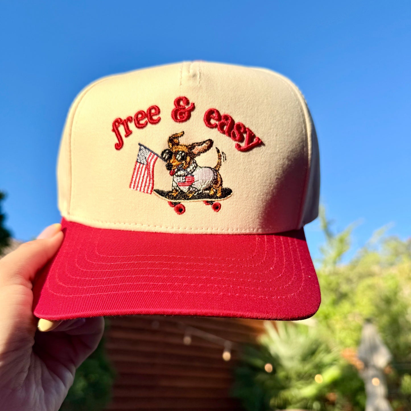 Beige and red cap with 'free & easy' text and dog graphic held against a blue sky.