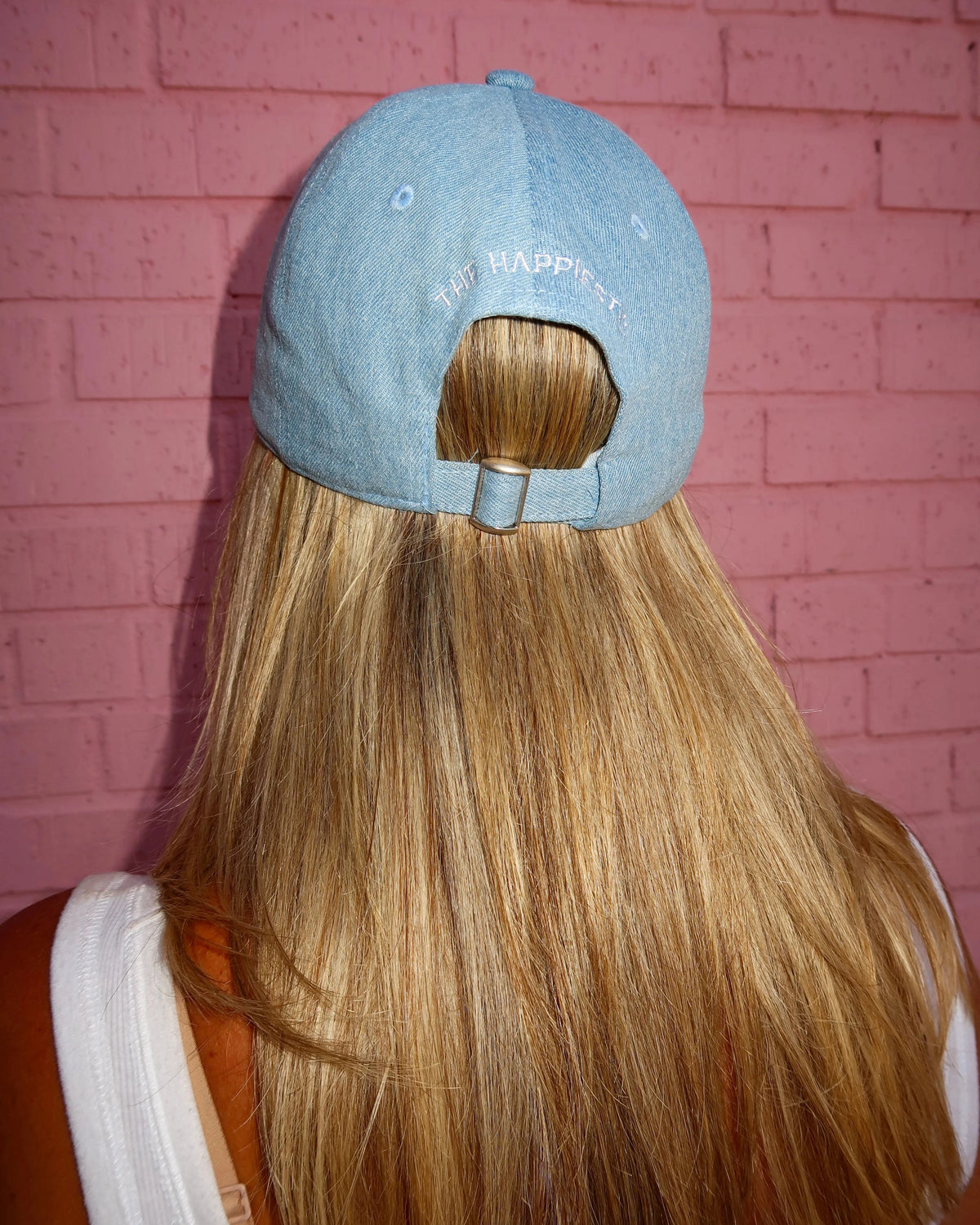 A person viewed from behind wearing a light blue denim baseball cap with 'HAPPY' embroidered on the back, shown against a pink brick wall.