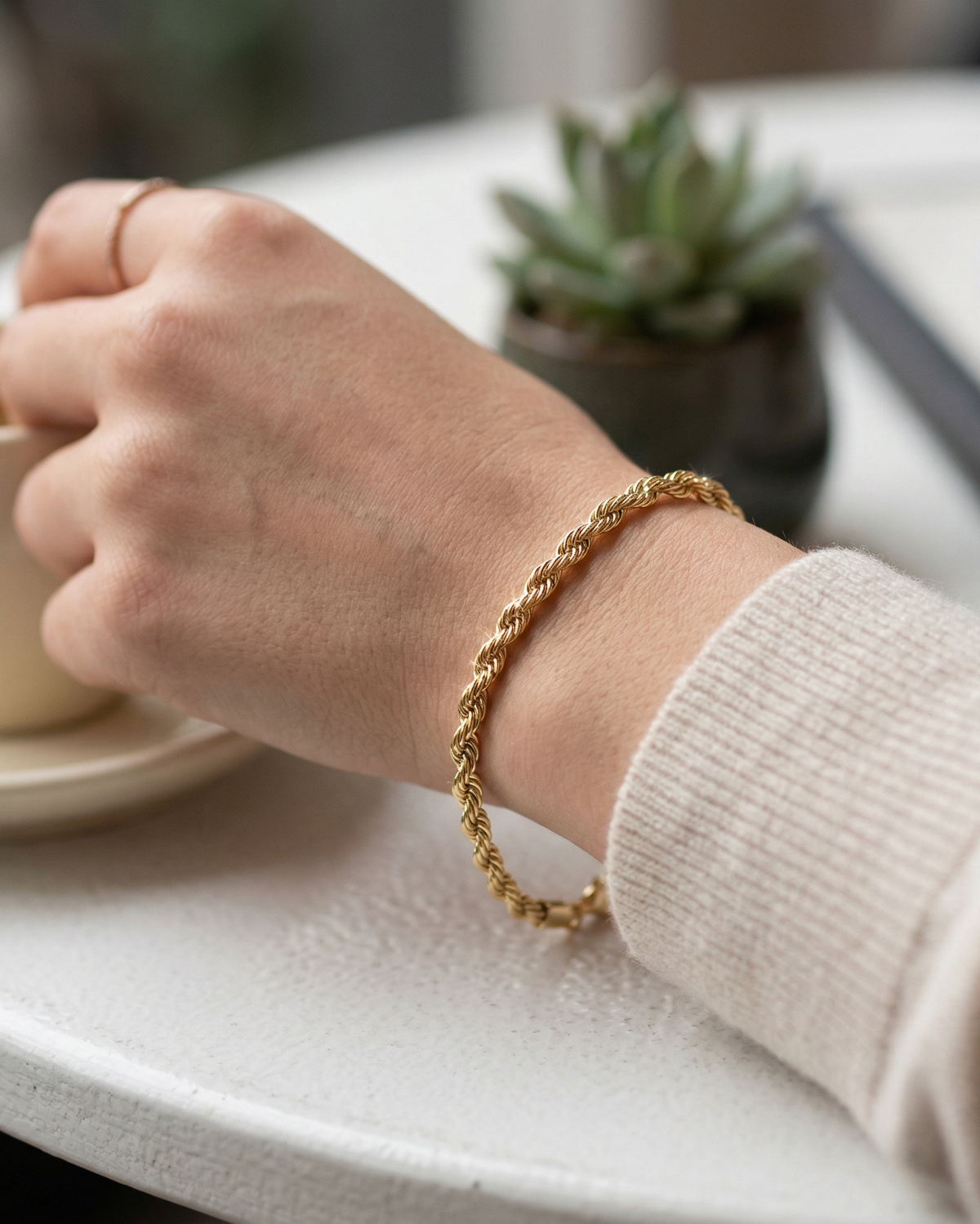 Hand wearing a gold bracelet holding a cup on a table with a plant in the background