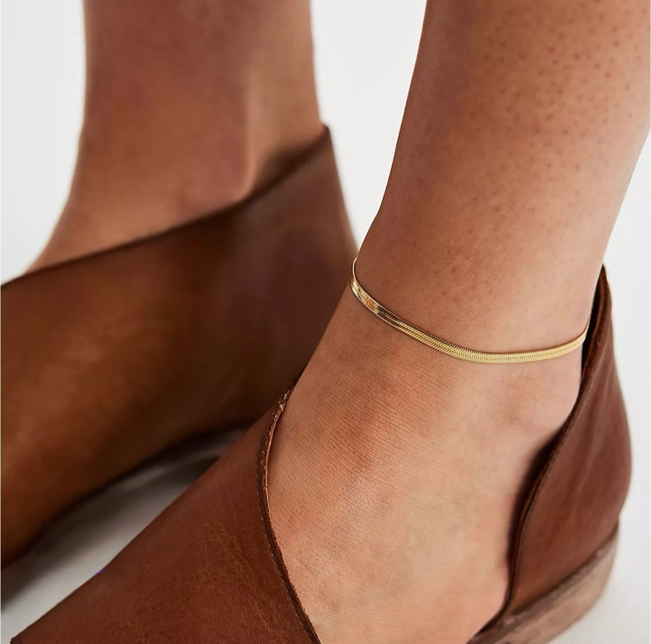 Brown suede shoes with a gold anklet on a neutral background