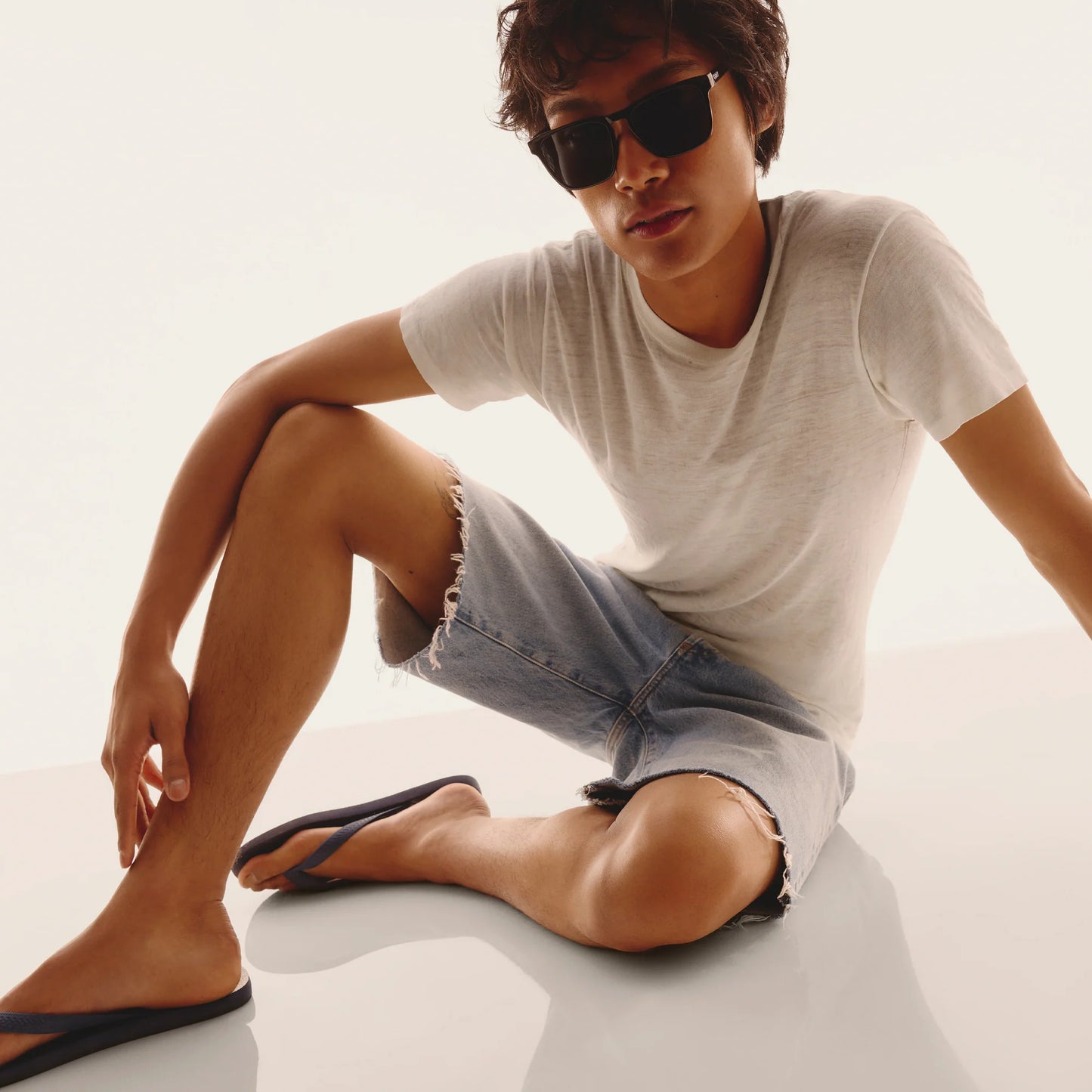Man wearing sunglasses and casual clothing sitting on a white surface.