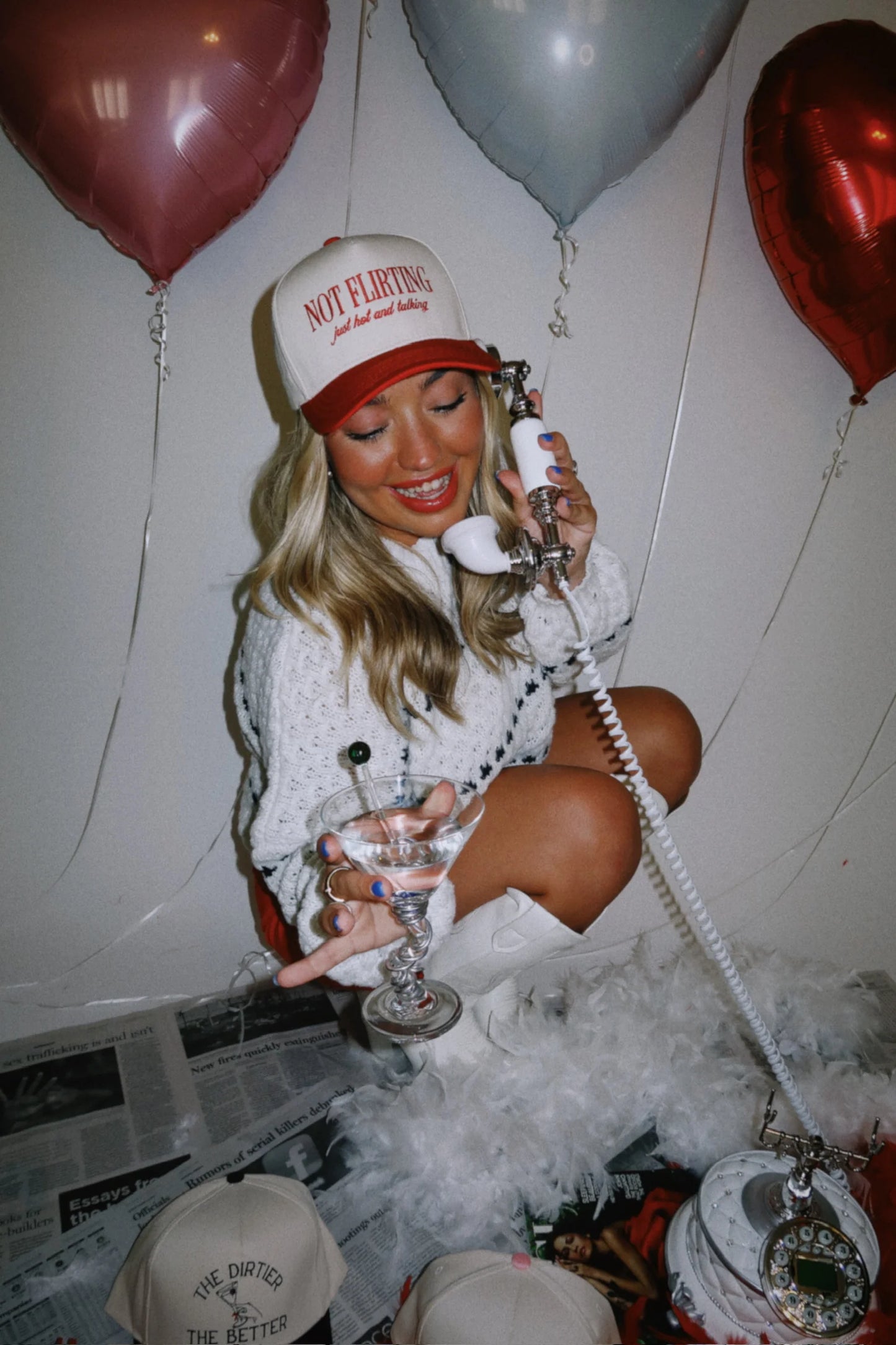 Person sitting on the floor with balloons and a phone, wearing a 'Not Flirting' cap.