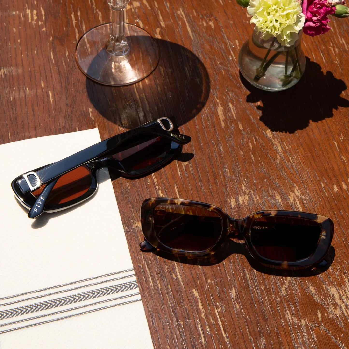 Two pairs of sunglasses on a wooden table with a glass and flowers in the background.