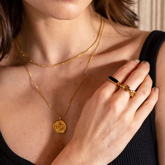 Close-up of a person wearing a gold necklace and ring, with a neutral background.