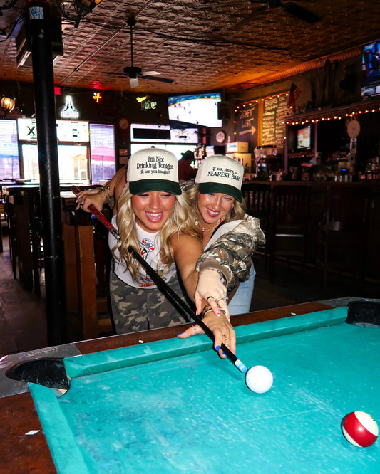 Two women smiling and playing pool at a bar, wearing matching beige and green baseball caps that read 'I'm So I Can Hardest Way' and 'I'm So I Can't Hard Way'.