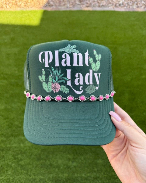 A dark green trucker hat with 'Plant Lady' embroidered in white, decorated with green cactus and pink flower designs, and accented with a pink rhinestone band. The hat is held by a hand against a background of green artificial turf.
