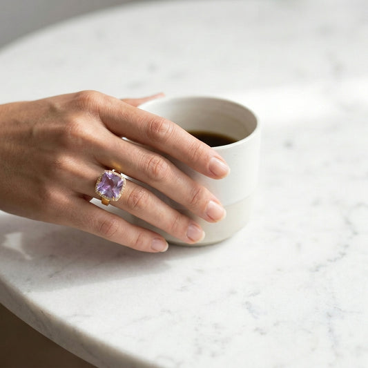 Hand wearing a ring with a purple gemstone holding a white mug on a light surface