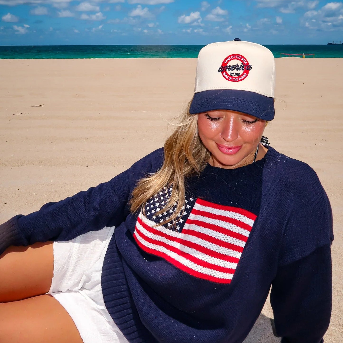 Woman sitting on a beach wearing a navy sweater with an American flag design and a white cap.