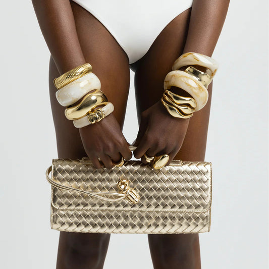 Gold handbag held by a person wearing gold and white bangles on a light gray background