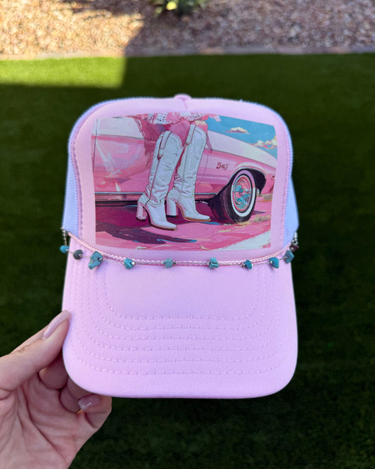 A hand holds a light pink trucker hat with a colorful graphic print of a pink car and white boots, decorated with a beaded rope across the front, set against a background of green grass and a gravel walkway.