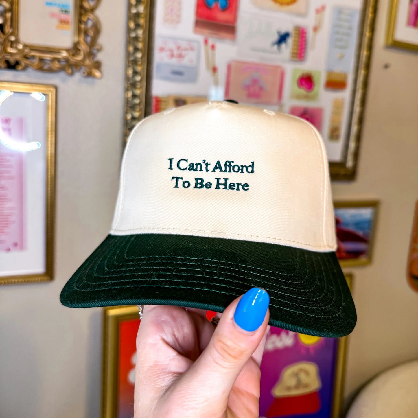 Trucker hat with 'I Can't Afford To Be Here' text held in front of framed pictures on a wall.