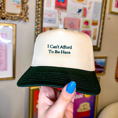 Trucker hat with 'I Can't Afford To Be Here' text held in front of framed pictures on a wall.