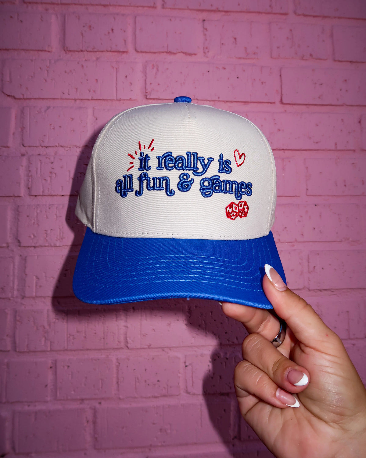 A white and blue snapback cap held by a hand against a pink brick wall. The front of the cap features embroidered blue text reading 'I really is all fun & games' with small red hearts and a red heart patch.