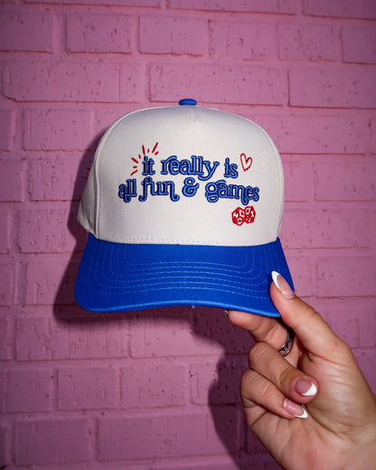 A white and blue snapback cap held by a hand against a pink brick wall. The front of the cap features embroidered blue text reading 'I really is all fun & games' with small red hearts and a red heart patch.