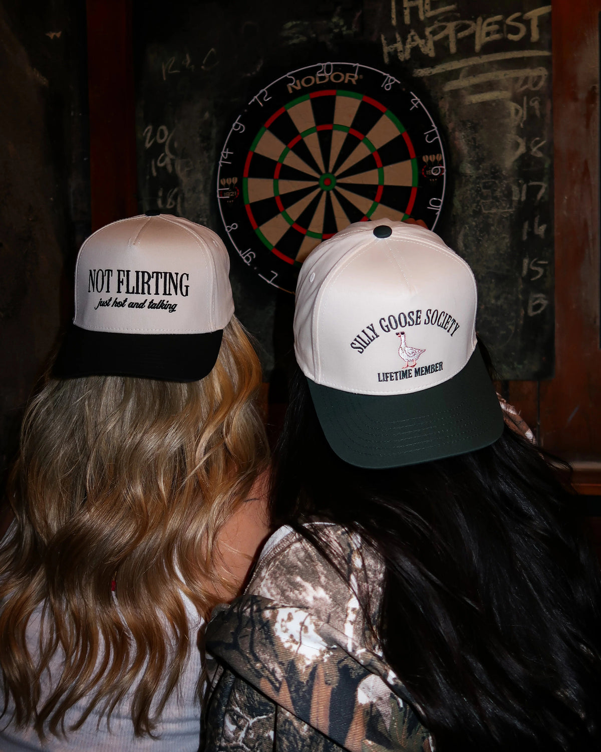 Two people wearing novelty hats in front of a dartboard with 'The Happiest' text on a wooden wall.