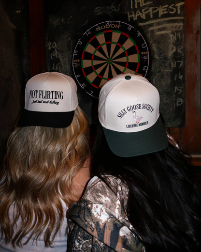 Two people wearing novelty hats in front of a dartboard with 'The Happiest' text on a wooden wall.