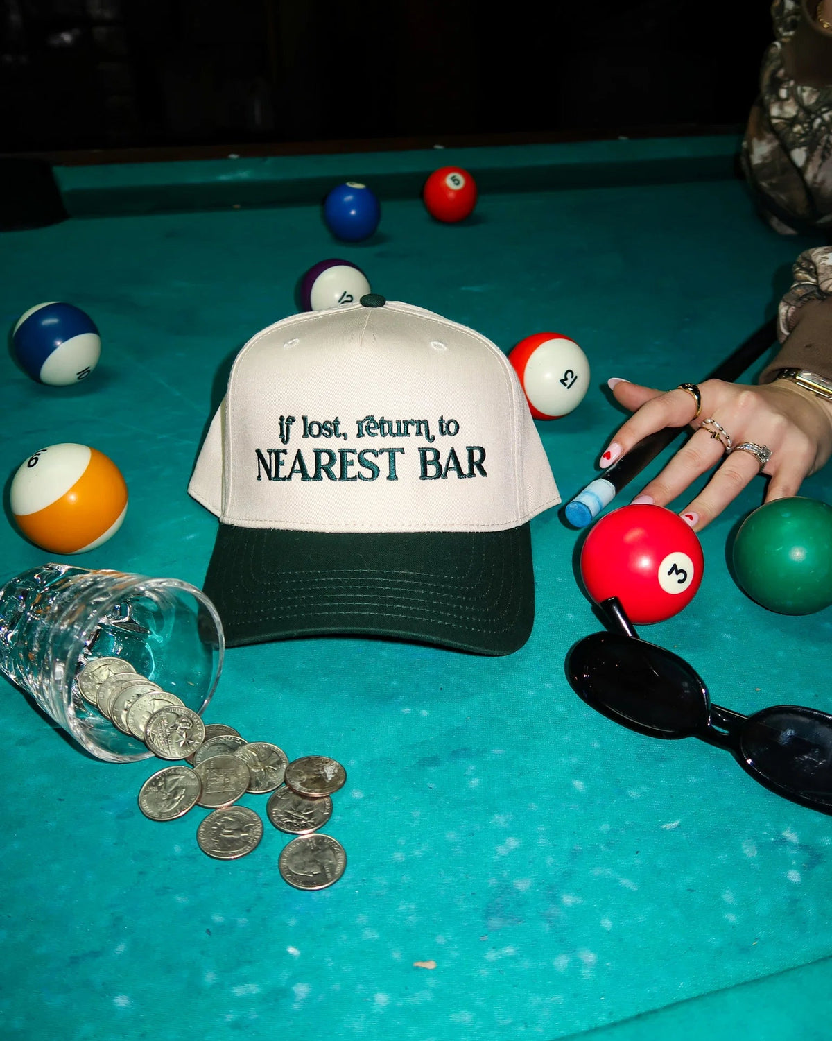 Cap with 'If lost, return to nearest bar' text on a pool table with pool balls and coins.
