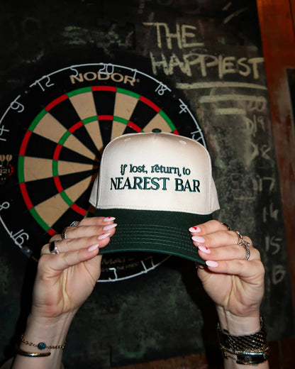 Person holding a cap with 'If lost, return to nearest bar' text in front of a dartboard.