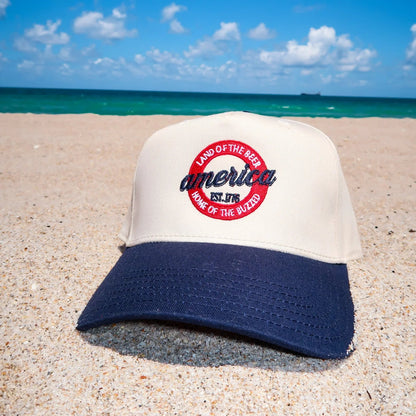 Cap with 'Land of the Free because of the Brave' text on a beach with ocean and sky in the background