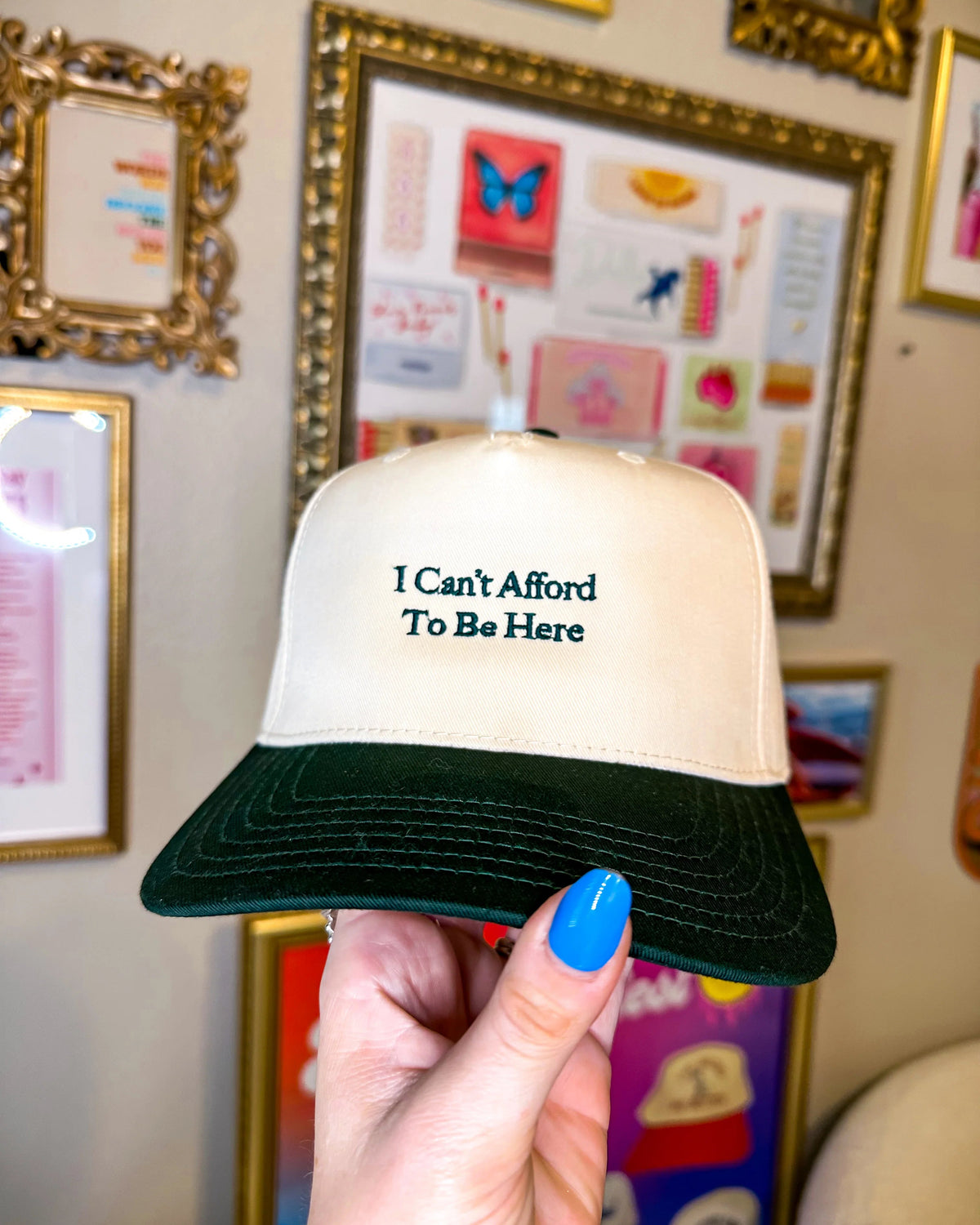 Trucker hat with 'I Can't Afford To Be Here' text held in front of framed pictures on a wall.