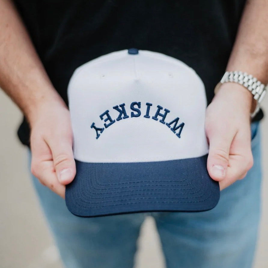 Person holding a white and navy blue cap with 'WHISKEY' printed on it.