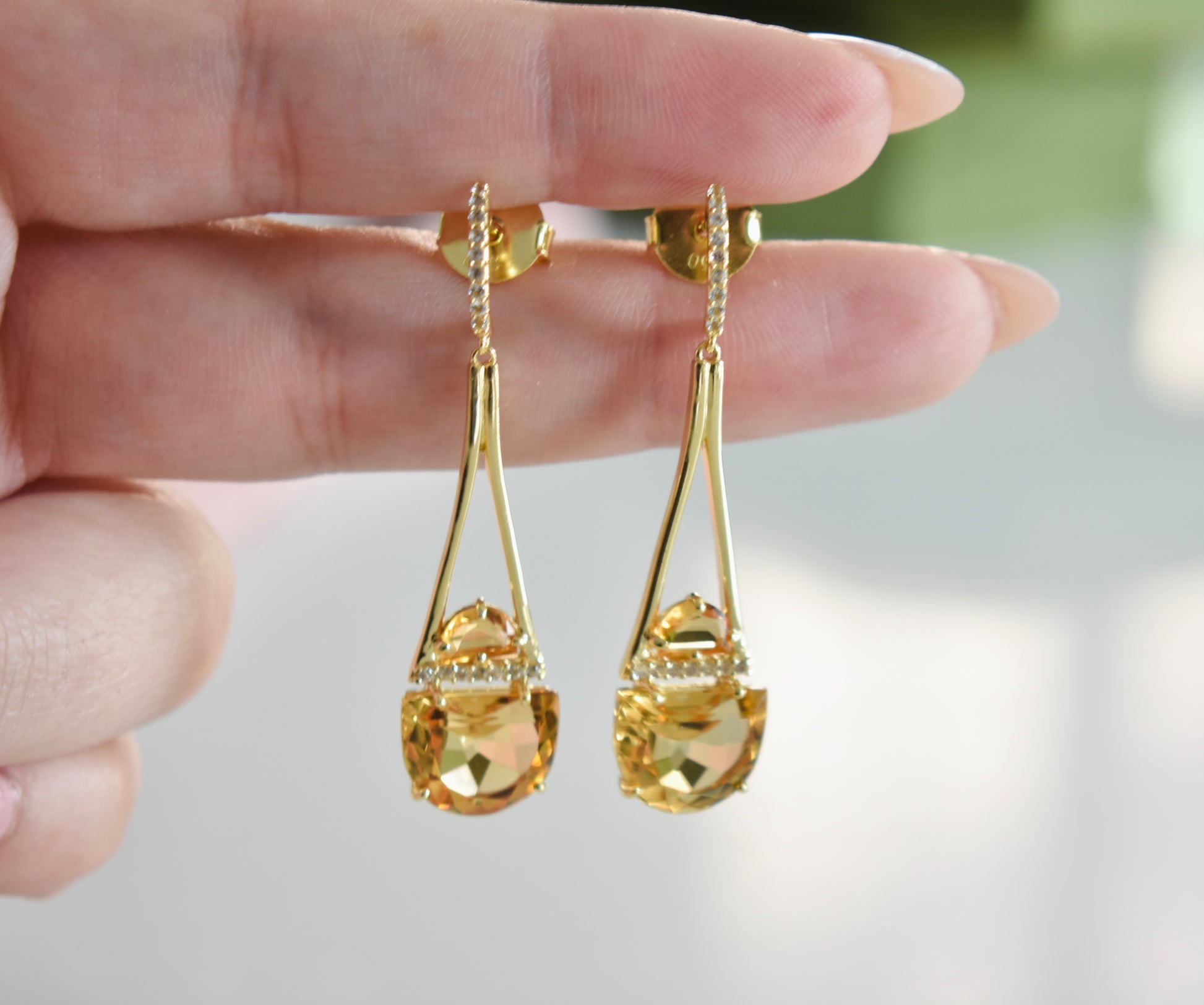 Gold earrings with clear stones held between fingers against a blurred background