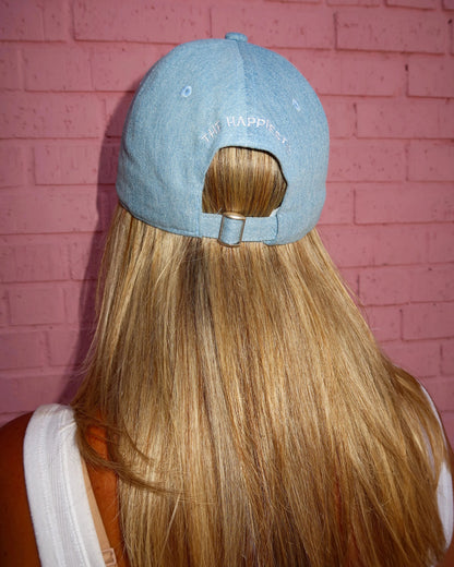 A person viewed from behind wearing a light blue denim baseball cap with 'HAPPY' embroidered on the back, shown against a pink brick wall.