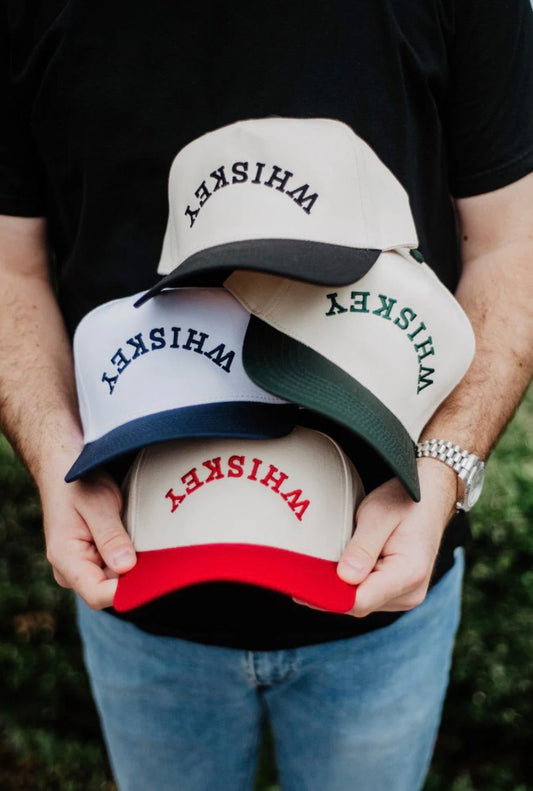 Four baseball caps with 'WHISKEY' text held by a person outdoors.