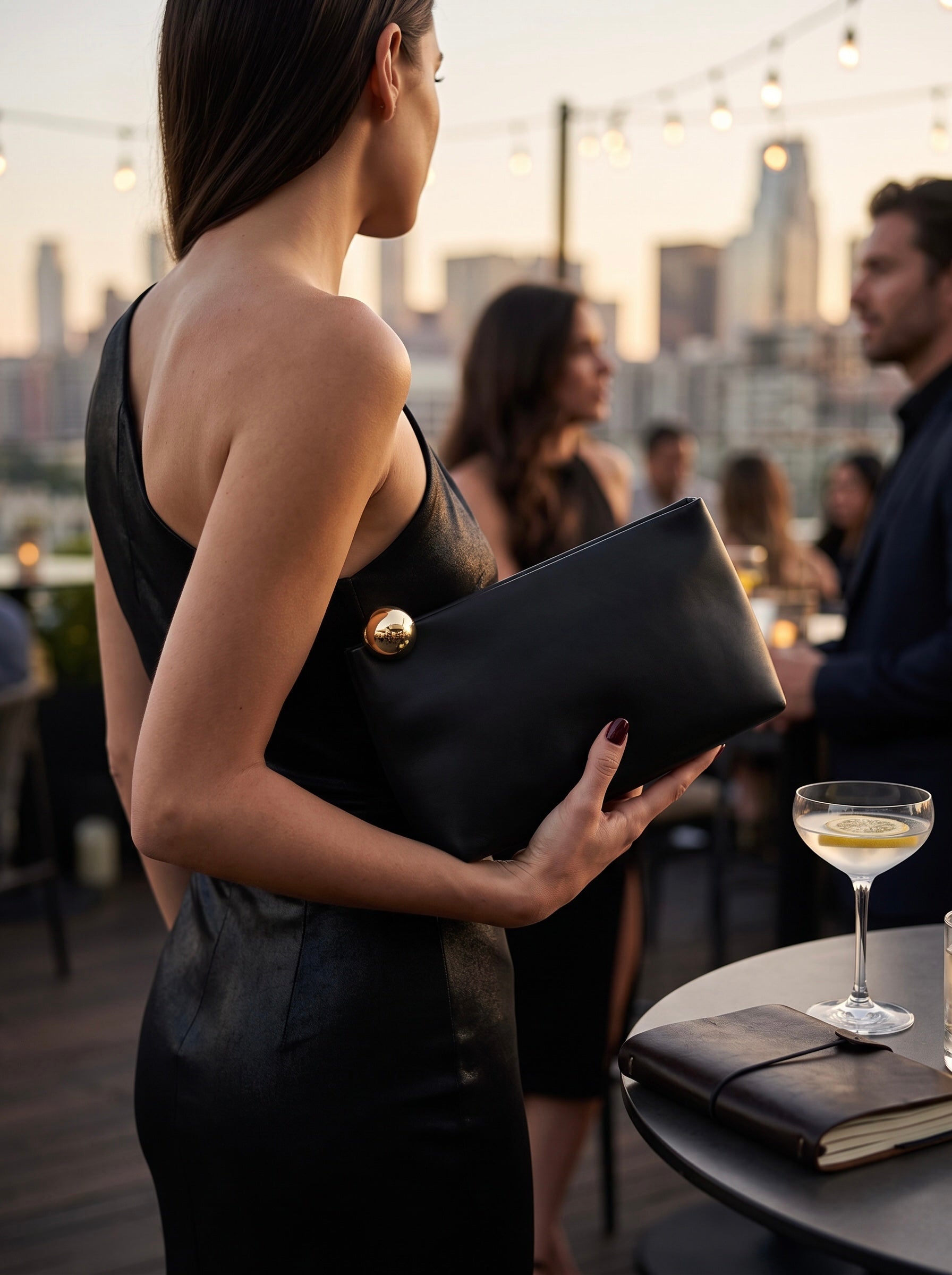 A woman in a black dress holds a black leather clutch with a gold button clasp on a rooftop terrace at dusk, with city buildings and other guests blurred in the background.