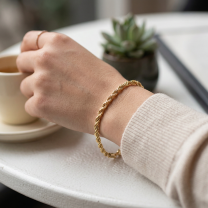 Hand wearing a gold bracelet holding a cup on a table with a plant in the background