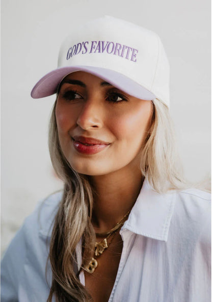 Woman wearing a white cap with 'GOD'S FAVORITE' text on a plain background