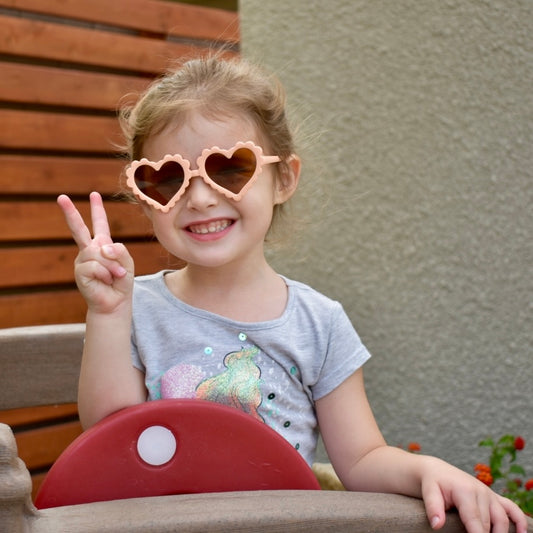 Child wearing heart-shaped sunglasses and making a peace sign outdoors.