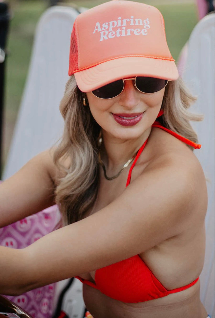 Woman wearing an 'Aspiring Retiree' cap and sunglasses, sitting on a beach chair.