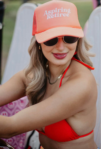 Woman wearing an 'Aspiring Retiree' cap and sunglasses, sitting on a beach chair.