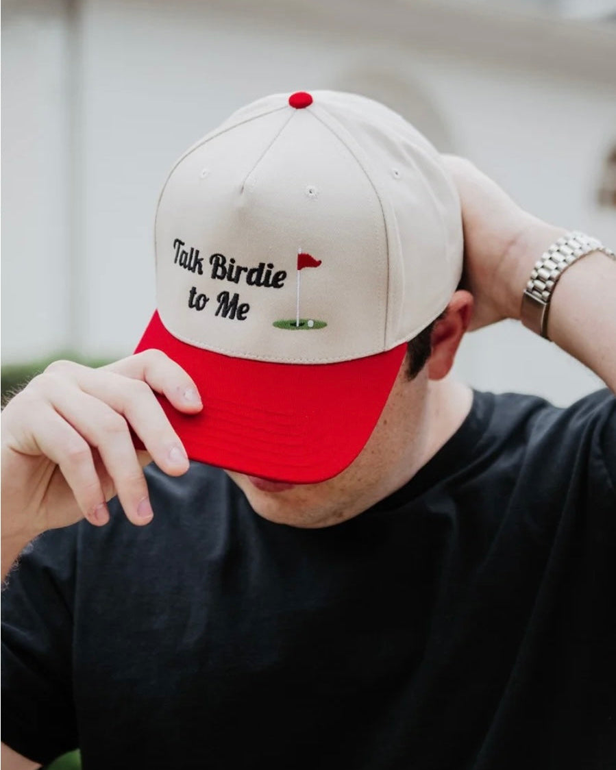 A man wearing a beige and red baseball cap with the text 'Talk Birdie to Me' and a small golf hole graphic on the front, holding the brim down with one hand.