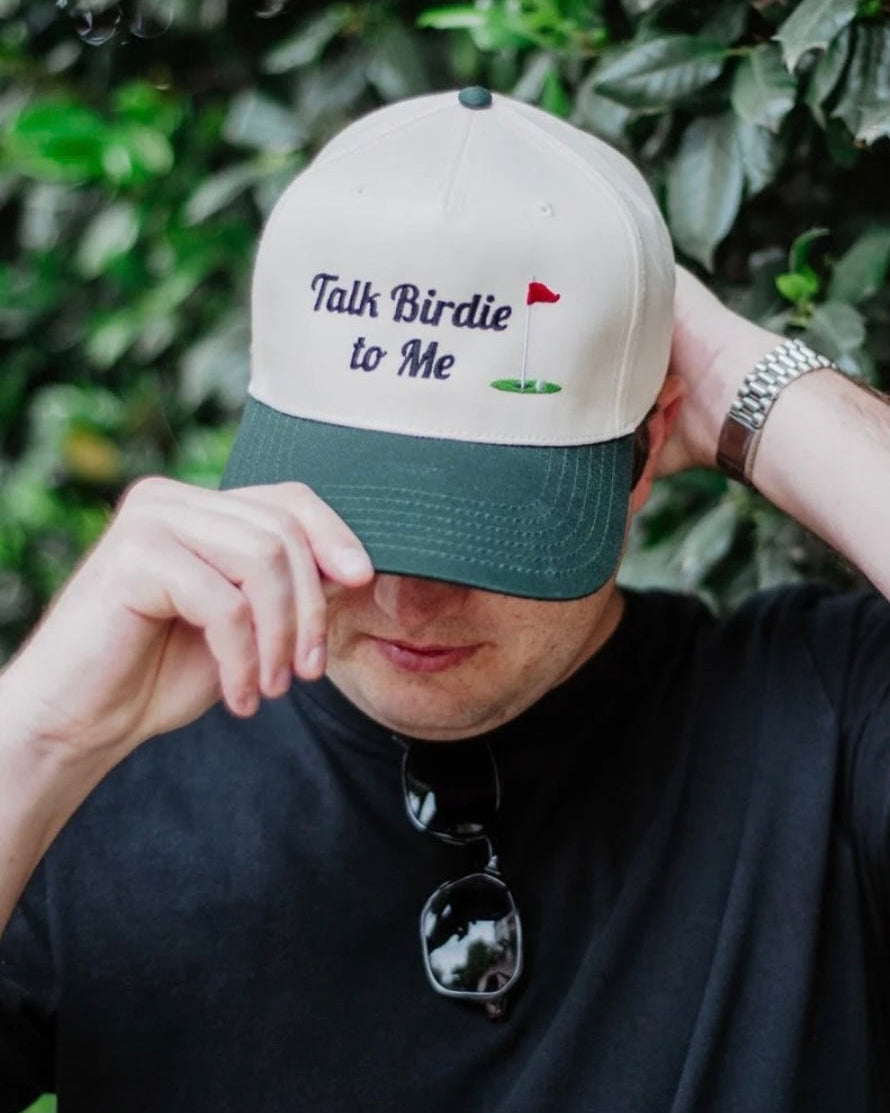 A man wearing a beige and dark green baseball cap with the embroidered text 'Talk Birdie to Me' and a small golf hole graphic, posed in front of a green leafy background.