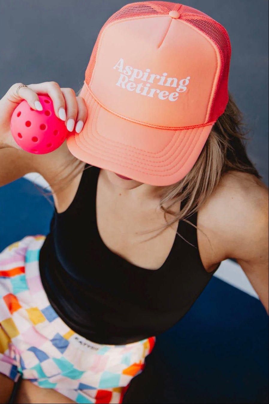 Person wearing a coral cap with 'Aspiring Retiree' text, holding a pink ball against a dark background.
