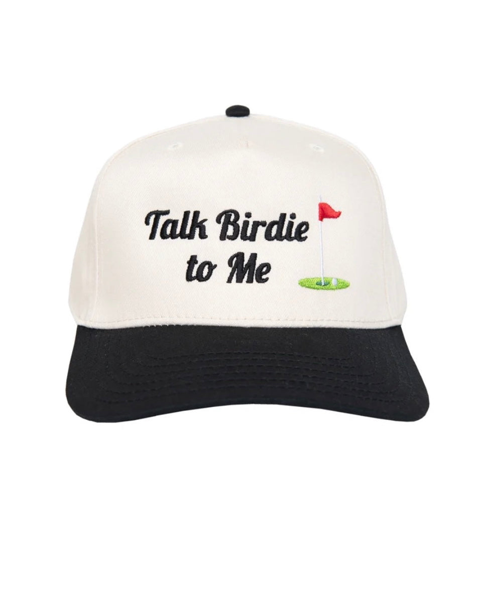A baseball cap with an off-white crown and a black curved brim. The front of the cap features the embroidered text 'Talk Birdie to Me' in black, alongside a small graphic of a green golf hole with a red flag.
