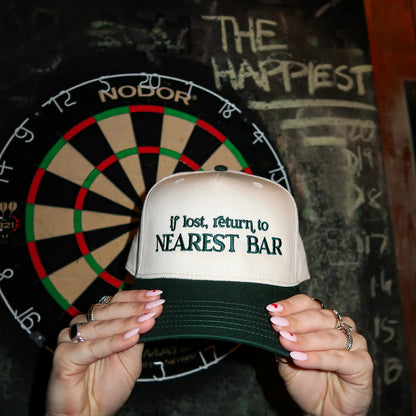 Person holding a cap with 'If lost, return to nearest bar' text in front of a dartboard.