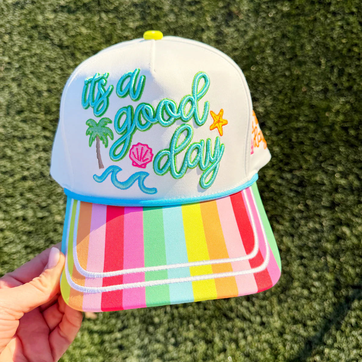 Colorful cap with 'It's a good day' text and palm tree design on a grass background