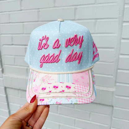 Cap with 'it's a very good day' text held against a white brick wall.