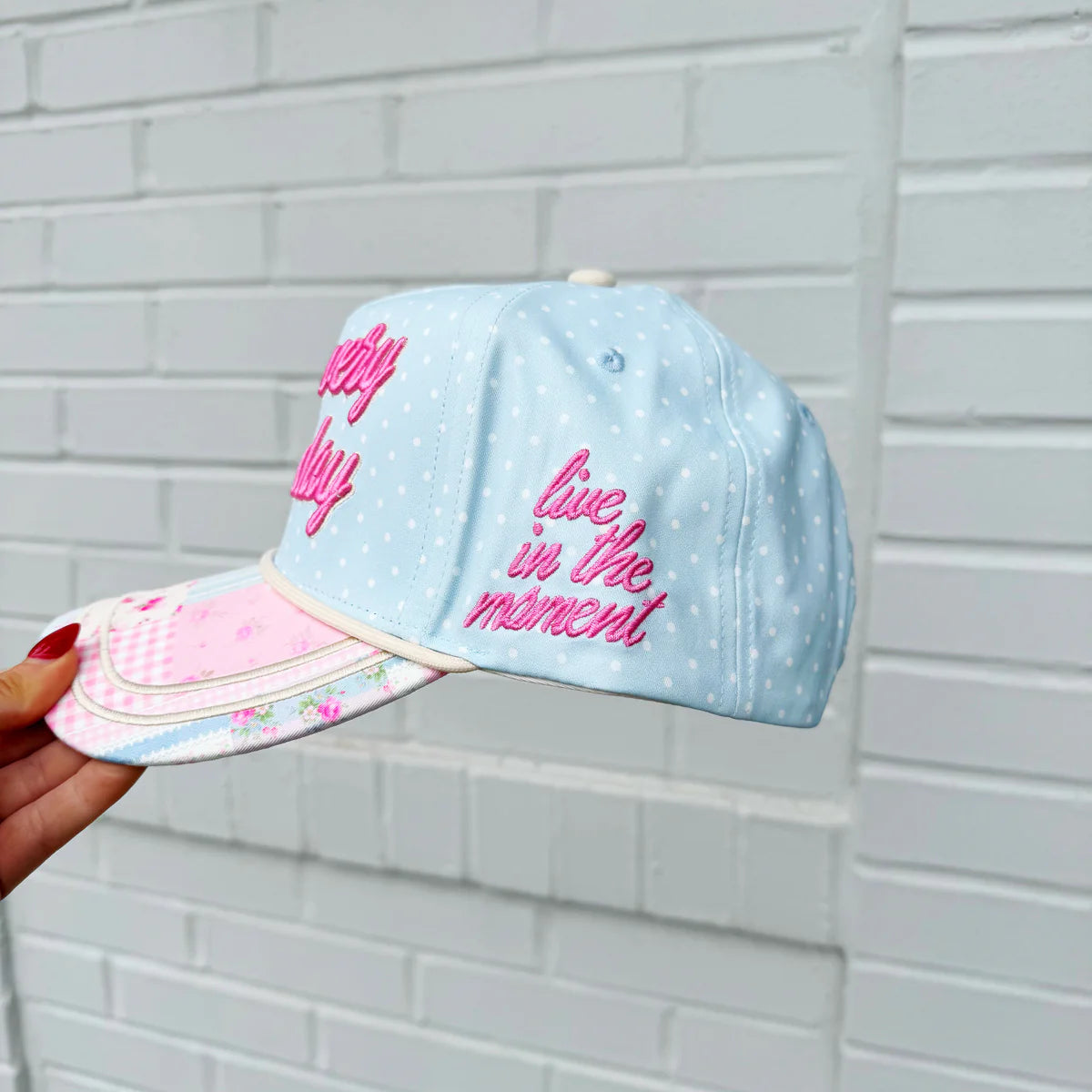 Light blue cap with pink text held against a white brick wall