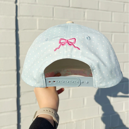 Light blue cap with pink bow design held against a white brick wall.