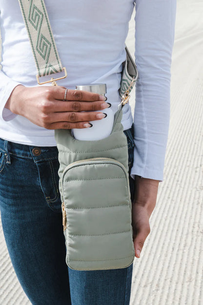 Person holding a cup with a green puffer bag over their shoulder on a light background