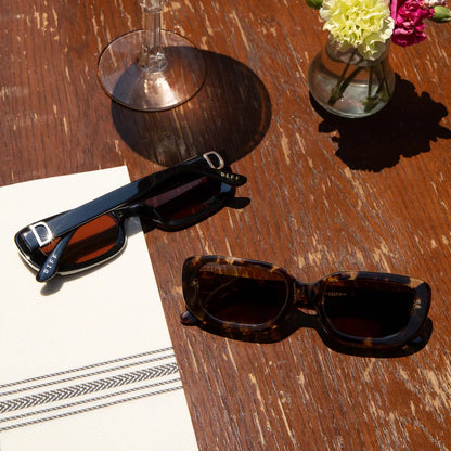Two pairs of sunglasses on a wooden table with a glass and flowers in the background.