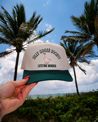Cap with 'Silly Goose Society Lifetime Member' text held in front of palm trees and blue sky.