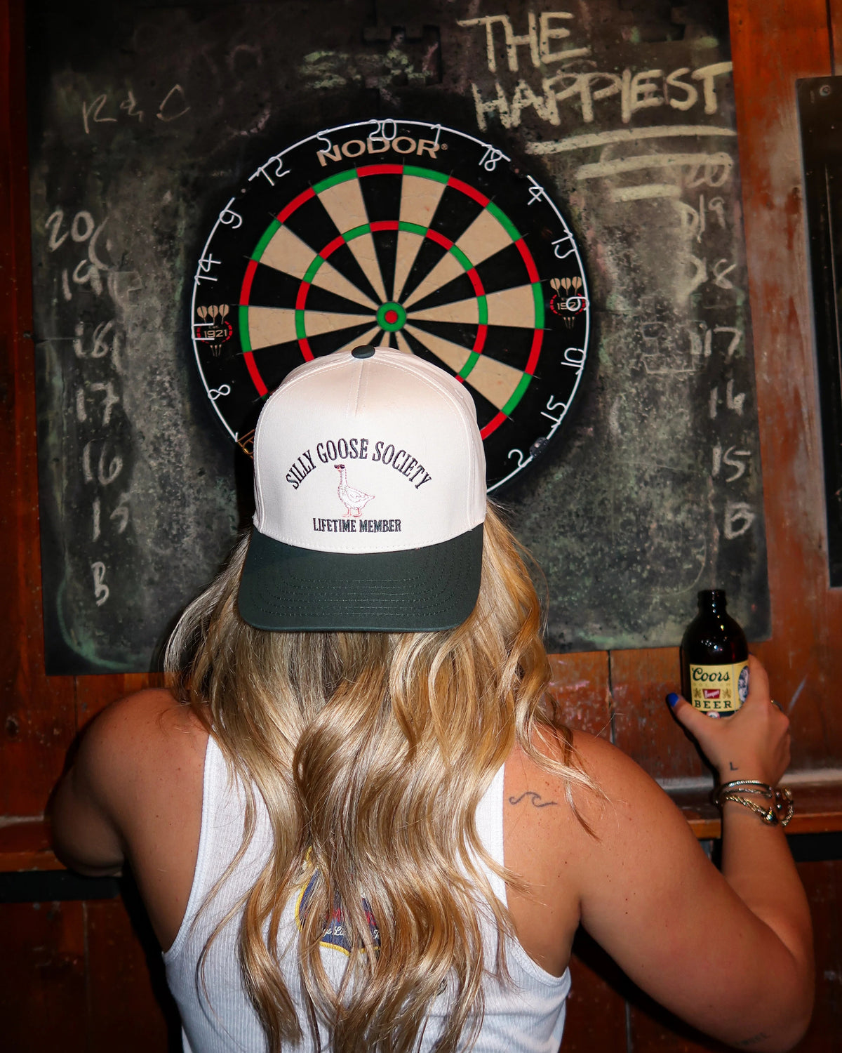 Person wearing a cap with 'Gulf Oil Society' branding, holding a bottle, in front of a dartboard.