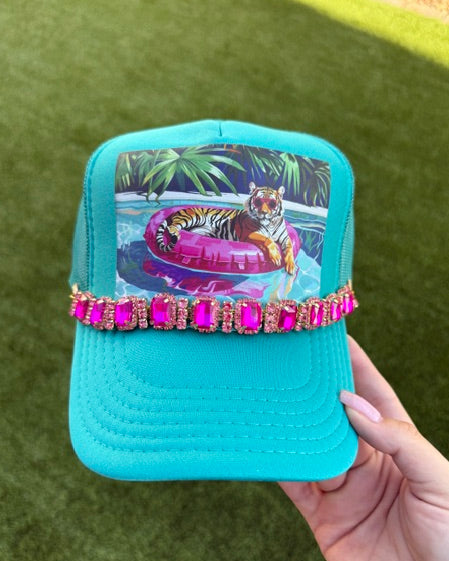 A turquoise blue trucker hat with a pink rhinestone band and a colorful graphic of a tiger on a pool float, held by a hand against a green grass background.