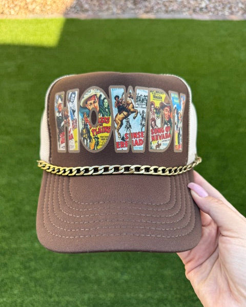 A hand holds a brown and beige trucker hat decorated with a gold chain across the brim and a colorful 'Cowboy' graphic on the front panel, set against a green artificial turf background.