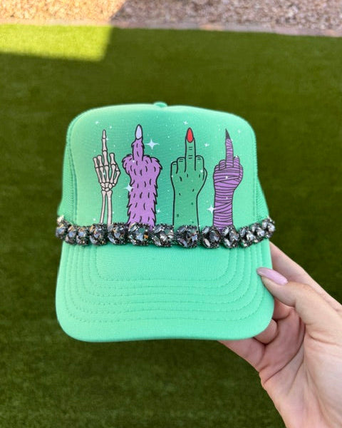 A mint green trucker hat held by a hand, featuring a graphic of four stylized hands with purple, pink, and black designs, and a row of black rhinestones across the brim, set against an outdoor grass background.